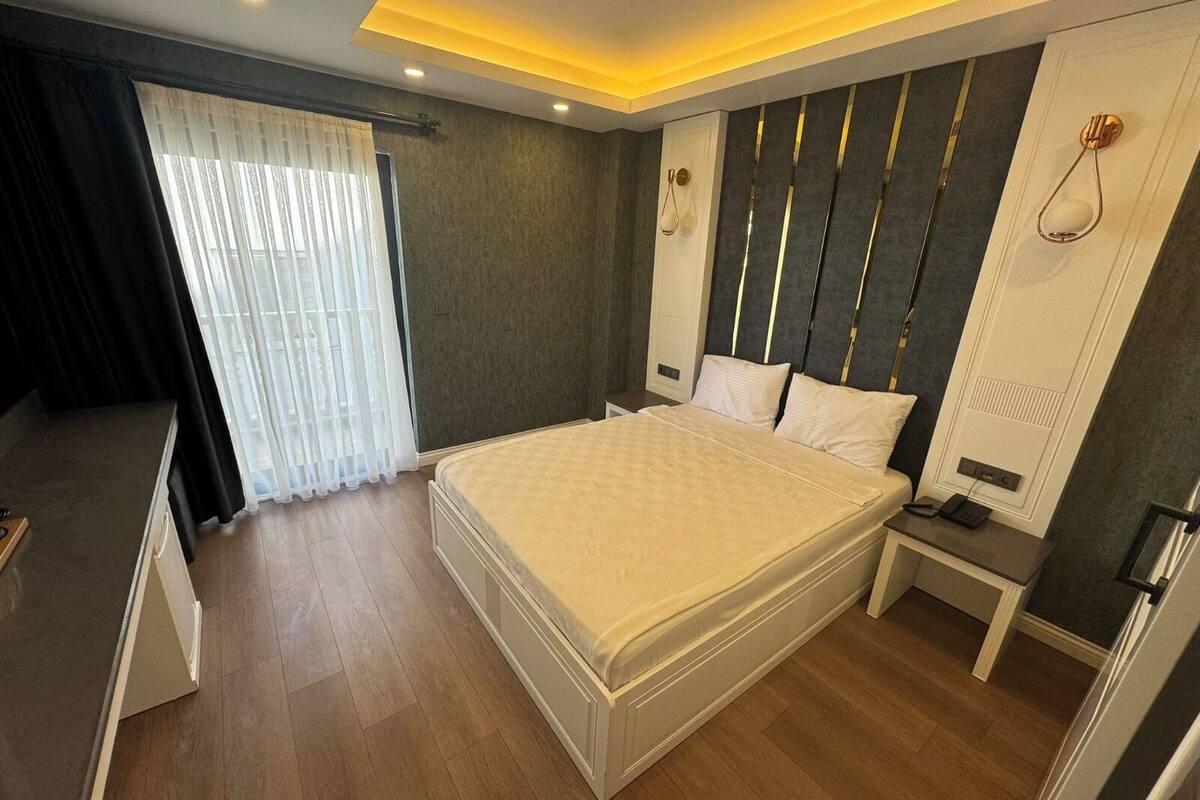 A modern bedroom with a large bed, bedside tables, a built-in wardrobe, and a window with sheer curtains.
