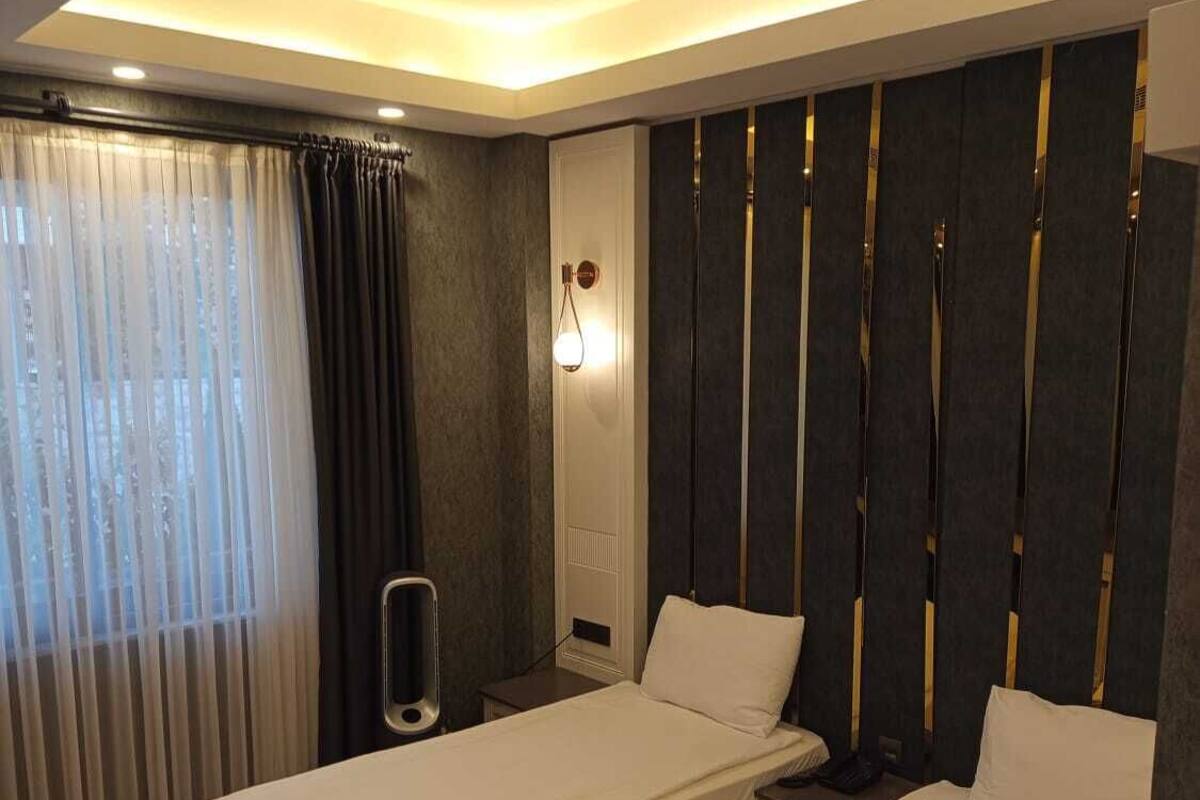 A hotel room with two beds, a large window with sheer curtains, a wall-mounted light fixture, and a dark-colored headboard.