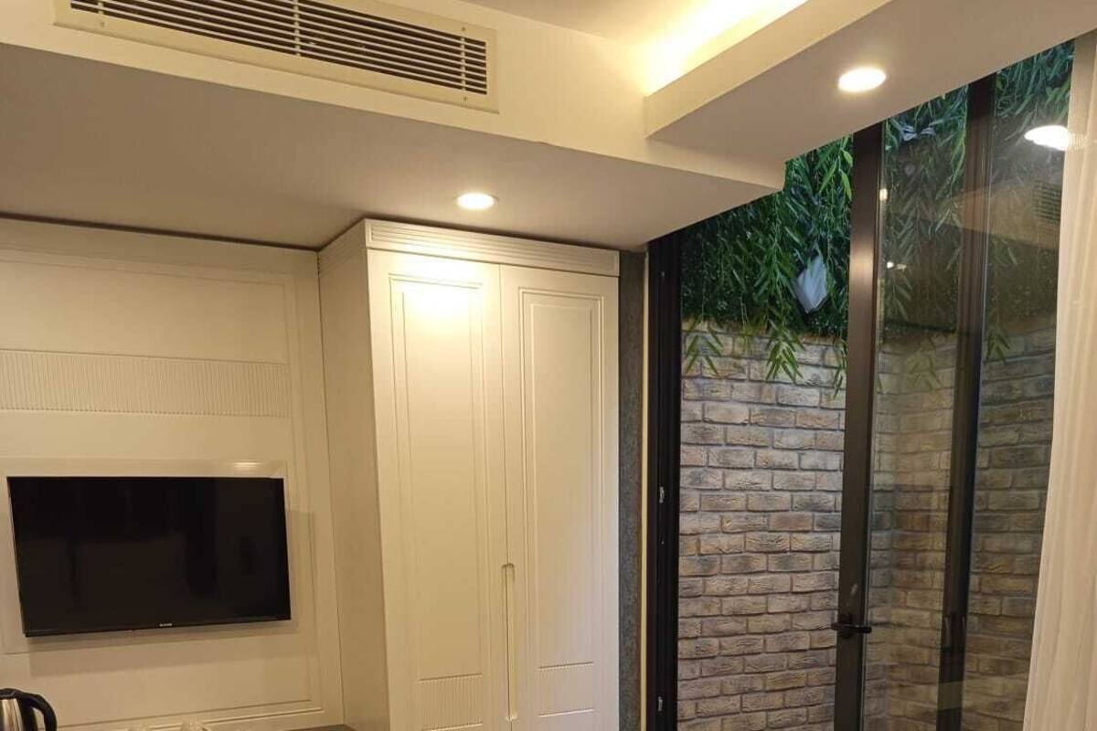A bedroom with a bed, a TV, a wardrobe, and a sliding glass door leading to a balcony with a brick wall and greenery.