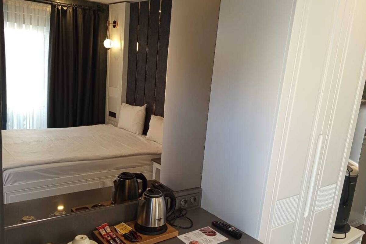 A hotel room with a bed, a small kitchenette, a desk with a kettle, cups, and a remote control.