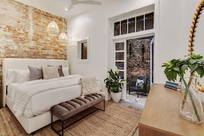 Une chambre avec un mur en briques, un lit blanc, un banc, une plante en pot et une table en bois sur laquelle est posé un vase.