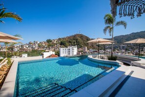 Una piscina con un diseño geométrico de azulejos, rodeada de sillones de descanso y sombrillas, con vistas a un pueblo en la ladera.