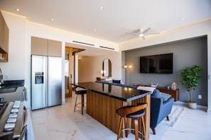 A modern kitchen with a central island, bar stools, a dining area with a sofa, and a flat-screen TV mounted on the wall.