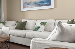 A modern living room with a beige sofa, green leaf-patterned pillows, and a framed landscape painting on the wall.