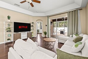 A modern living room with a flat-screen TV, white sofas, a coffee table, and a sliding glass door leading to an outdoor area.
