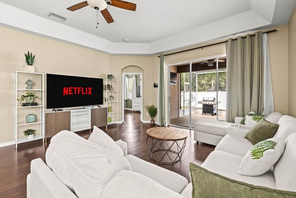 A modern living room with a flat-screen TV, white sofas, a coffee table, and a sliding glass door leading to an outdoor area.