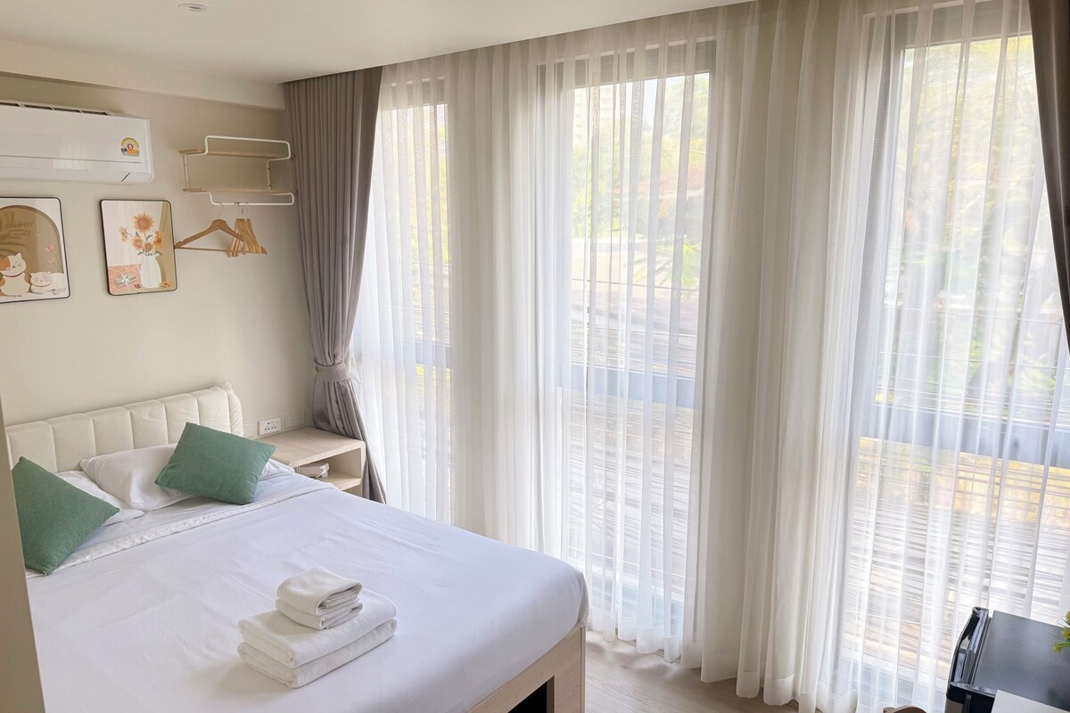 A hotel room with a bed, a bedside table, a chair, a desk, and a window with sheer curtains.