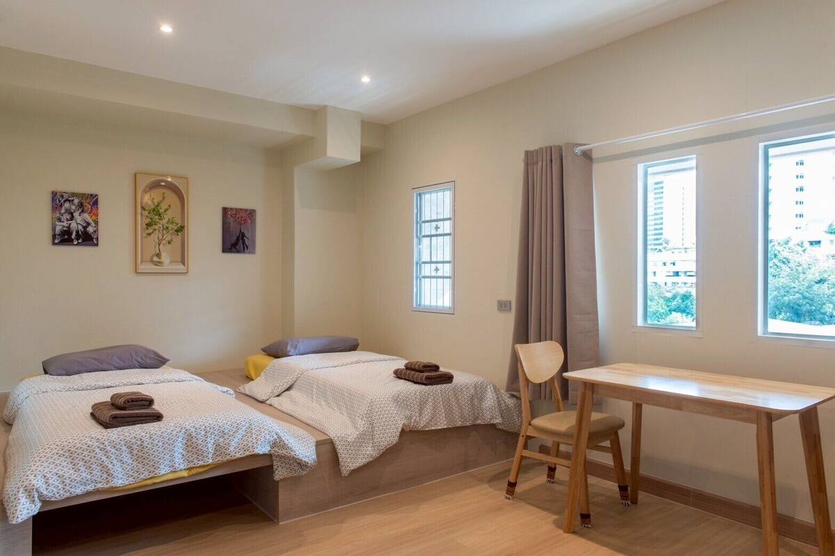 A bedroom with two beds, a wooden desk, and a chair. There are windows with curtains, and artwork on the walls.