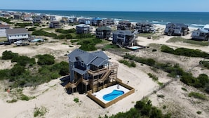 Un área residencial costera con varias casas, destacándose una casa de dos pisos con terraza de madera y piscina, y una playa al fondo.