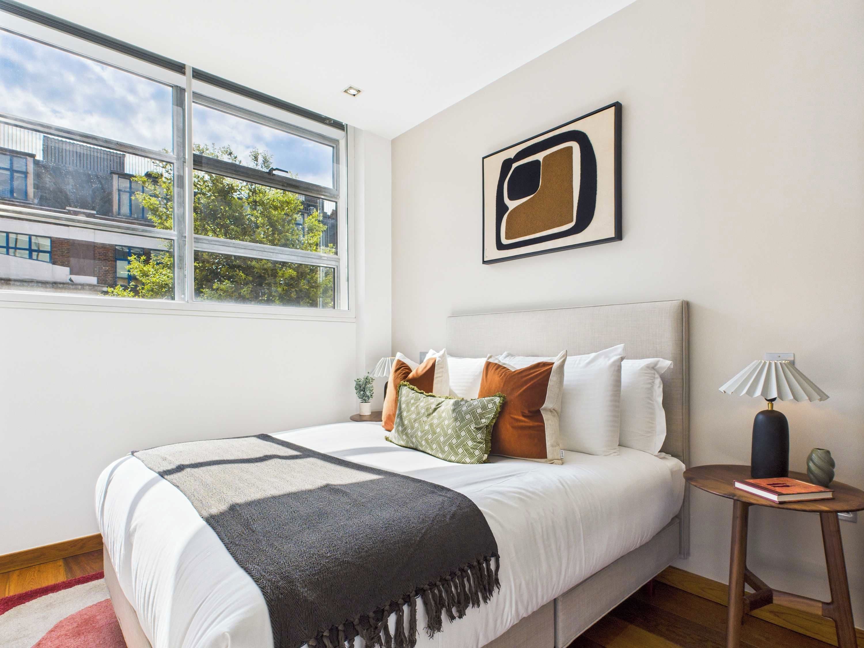 A modern bedroom with a large bed, a view of buildings outside, and a framed abstract painting on the wall.