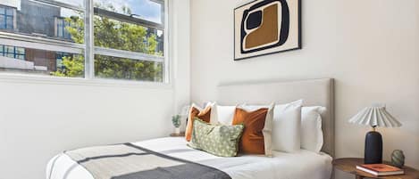 A modern bedroom with a large bed, a view of buildings outside, and a framed abstract painting on the wall.
