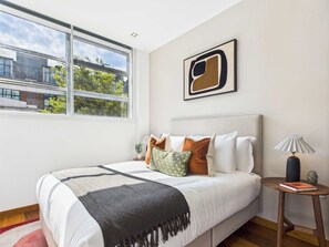 Une chambre à coucher moderne avec un grand lit, une vue sur les bâtiments environnants et un tableau abstrait encadré accroché au mur.