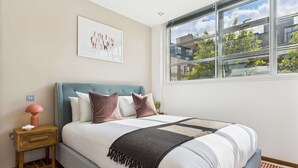 A bedroom with a bed, a nightstand, a lamp, a window with a view of buildings and trees, and a framed picture on the wall.
