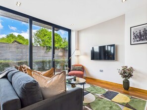 A modern living room with a large sofa, a red armchair, a glass coffee table, and a flat-screen TV mounted on the wall. - Bright Camden Town Apartment with Balcony (Greater London)