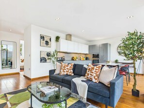 A modern living room with a dark blue sofa, a glass coffee table, and a view into the kitchen.