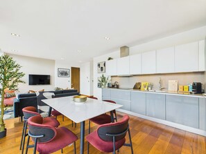 Una cocina moderna con armarios blancos, una zona de comedor con mesa blanca y sillas rojas, y una sala de estar con sofá y televisor.