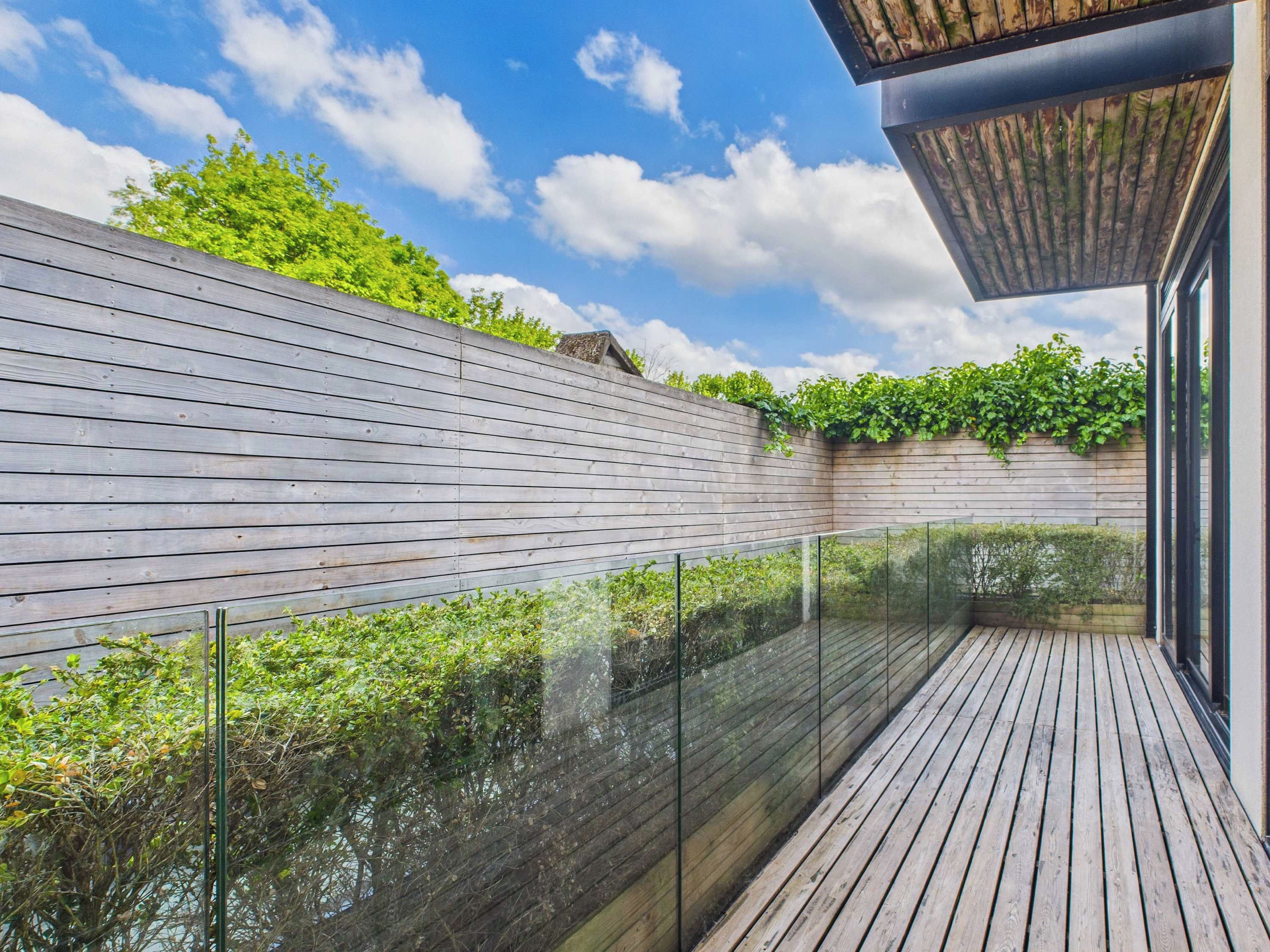 A modern outdoor deck with a glass railing, wooden flooring, and a wooden wall with greenery.