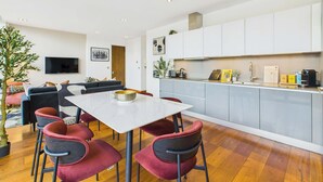 A modern kitchen with white cabinets, a dining area with a white table and red chairs, and a living area with a sofa and a TV.