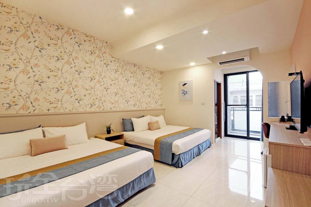 Ein Hotelzimmer mit zwei Betten, einem kleinen Nachttisch mit Pflanze, einem großen Fenster und einem Spiegel an der Wand.