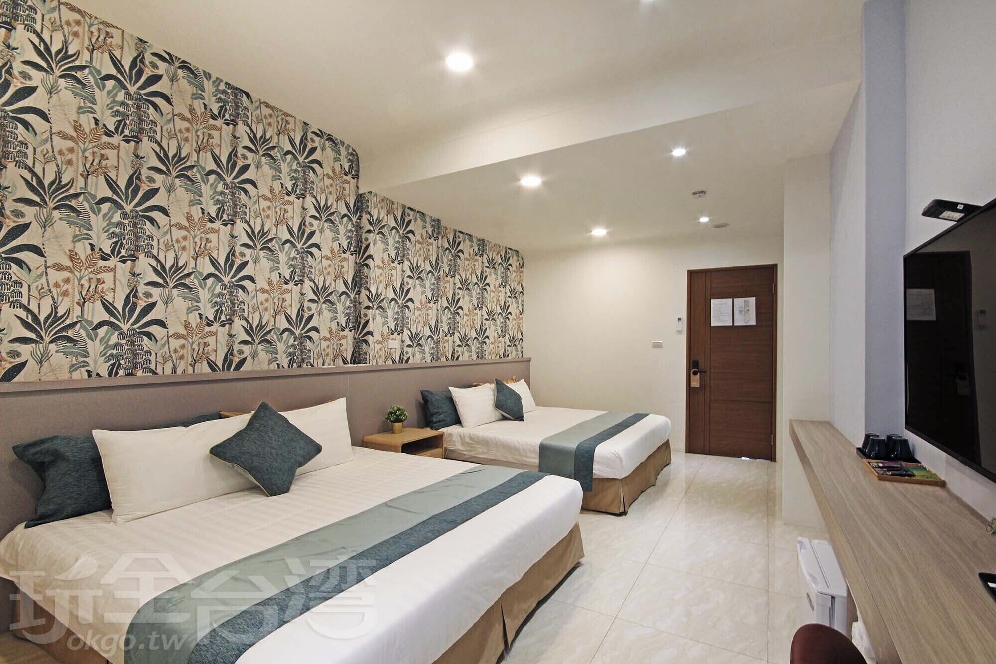 A hotel room with two beds, a large floral-patterned headboard, a small bedside table with a plant, and a TV on the right.