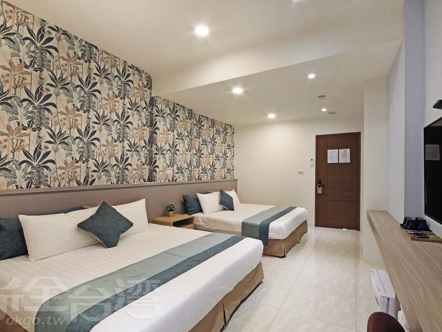 A hotel room with two beds, a large floral-patterned headboard, a small bedside table with a plant, and a TV on the right.