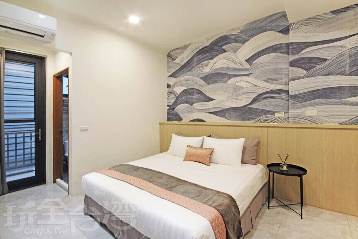 Ein Hotelzimmer mit einem großen Bett, einem Holzkopfende, einem an der Wand befestigten Kunstwerk, einem kleinen Nachttisch und einer Klimaanlage.