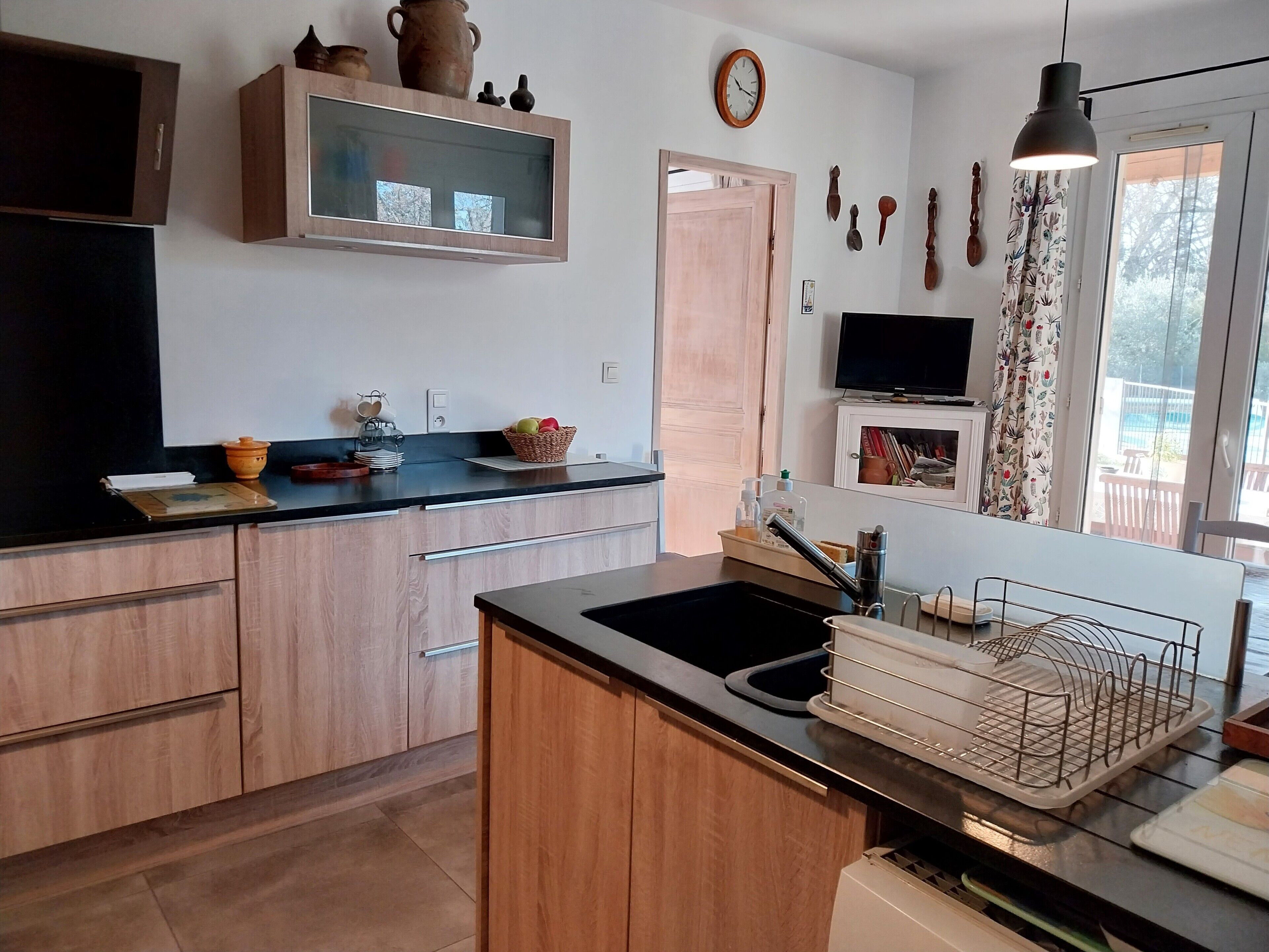 A modern kitchen with wooden cabinets, a black countertop, and a built-in sink. There is a television on the wall and a window with curtains.