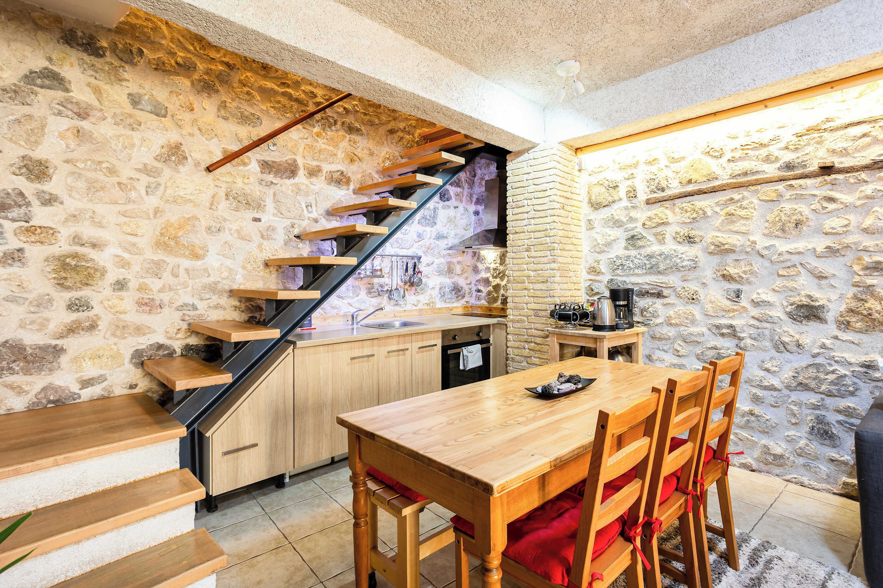 Une cuisine avec un mur en pierre, un escalier en bois et un coin repas comprenant une table et des chaises.