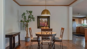 A dining area with a round table, four chairs, a potted plant, a framed painting, and a pendant light.