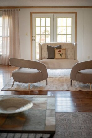 Een woonkamer met een bank en twee fauteuils, een groot raam met gordijnen, en een vloerkleed.