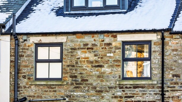 A stone building with a sloped roof, multiple windows, a glass door entrance, outdoor seating, and snow on the ground.