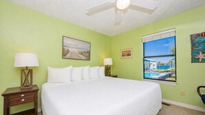 A hotel room with a large bed, two bedside tables with lamps, a ceiling fan, and a window with a view of a pool and buildings. - Sea Oats 105h 3 Bedroom Condo (Gulf Shores)