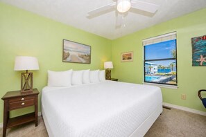 A hotel room with a large bed, two bedside tables with lamps, a ceiling fan, and a window with a view of a pool and buildings. - Sea Oats 105h 3 Bedroom Condo (Gulf Shores)