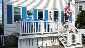 Een huis met twee verdiepingen, een witte gevel en blauwe luiken, een veranda met blauwe stoelen, en een Amerikaanse vlag.