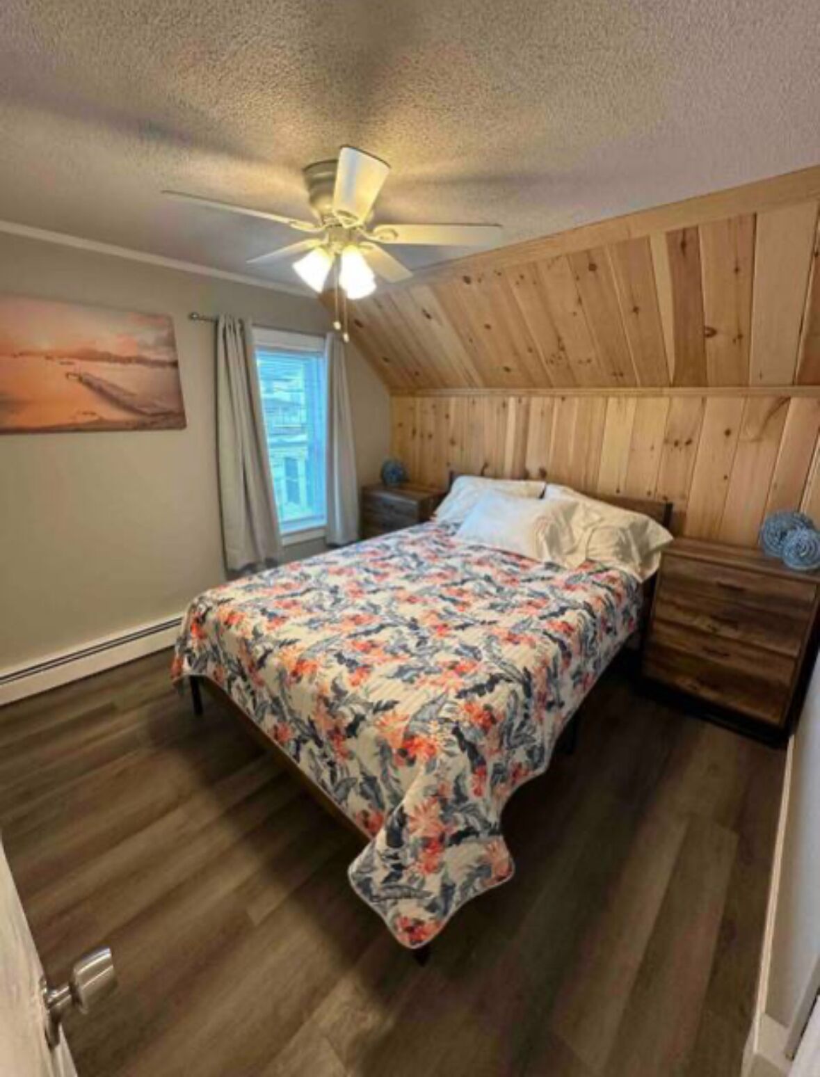 A bedroom with a wooden ceiling, a bed with a floral bedspread, a nightstand, and a ceiling fan.