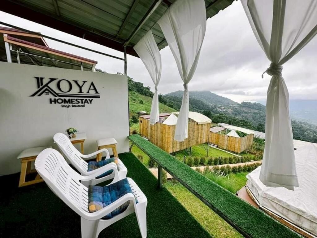 A covered outdoor seating area with a view of a mountainous landscape, a white wall with the text 'KOYA HOMESTAY,' and a wooden structure in the background.