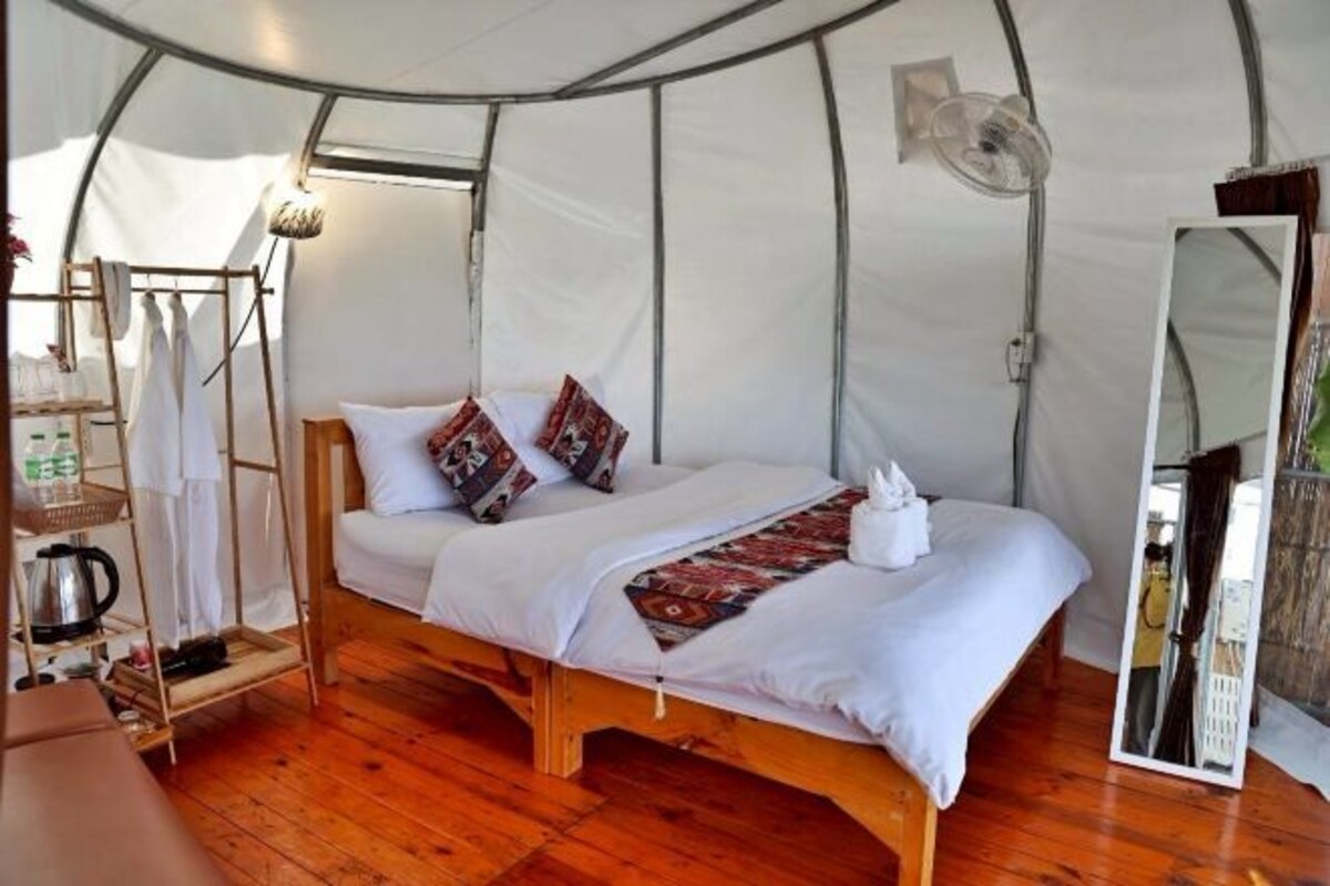 A tent with a bed, a wooden shelf with items, a mirror, and a hanging light fixture.