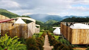 A rustic wooden cabin with a dirt path leading to it, surrounded by lush greenery and mountains in the background. - Koya Homestay (Mae Rim)