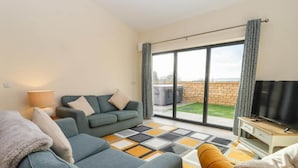 Un salón moderno con sofá, sillón y televisor, y grandes puertas correderas de vidrio a un jardín.