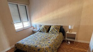 Een slaapkamer met een bed, twee nachtkastjes, een raam met zonwering en een houten visgraatvloer.
