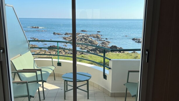 Een kamer met een schuifglazen deur die leidt naar een balkon met uitzicht op zee en rotsen.
