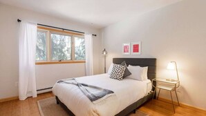Un dormitorio con cama, una mesita de noche con lámpara, una ventana con cortinas y dos cuadros en la pared.