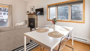 Una sala de estar moderna con una mesa de comedor blanca, sillas transparentes, un sofá, una chimenea, un televisor y un ventanal grande.
