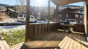 Una terraza de madera con una parrilla y una mesa, con vista a un estacionamiento con vehículos y casas al fondo.
