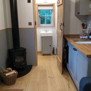 A narrow kitchen with a wood-burning stove, a window with a view, a sink, and cabinets. - Shepherd's Hut in Sussex With Fire Pit & Wildlife (Steyning)