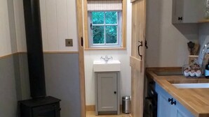 A narrow kitchen with a wood-burning stove, a window with a view of greenery, a sink, and cabinets. - Shepherd's Hut in Sussex With Fire Pit & Wildlife (Steyning)