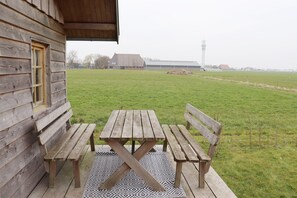 A wooden cabin with a picnic table and benches on a patterned mat, overlooking a grassy field and distant buildings. - Cottages in Friesland With Farm Experience (Tjerkgaast)
