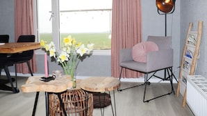 A room with a view of a green field, a table with a vase of flowers, a chair with a cushion, and a floor lamp. - Cottages in Friesland With Farm Experience (Tjerkgaast)