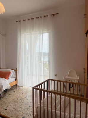 Un dormitorio con una cama, una cuna, un ventanal con cortinas translúcidas y una puerta.