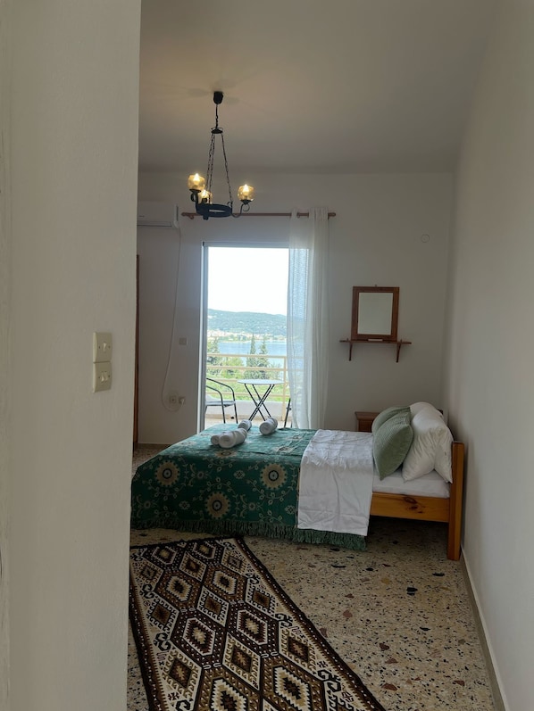 Un dormitorio con cama, una mesita, una silla, lámpara colgante, ventana con vistas y alfombra estampada.